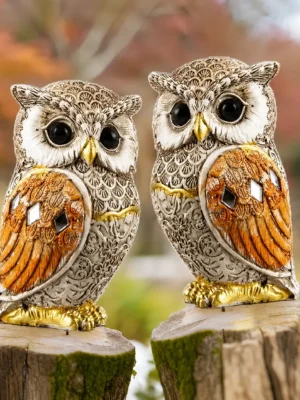 Pair of decorative owl statues with intricate feather detailing, golden accents, and reflective embellishments placed on a wooden surface.
